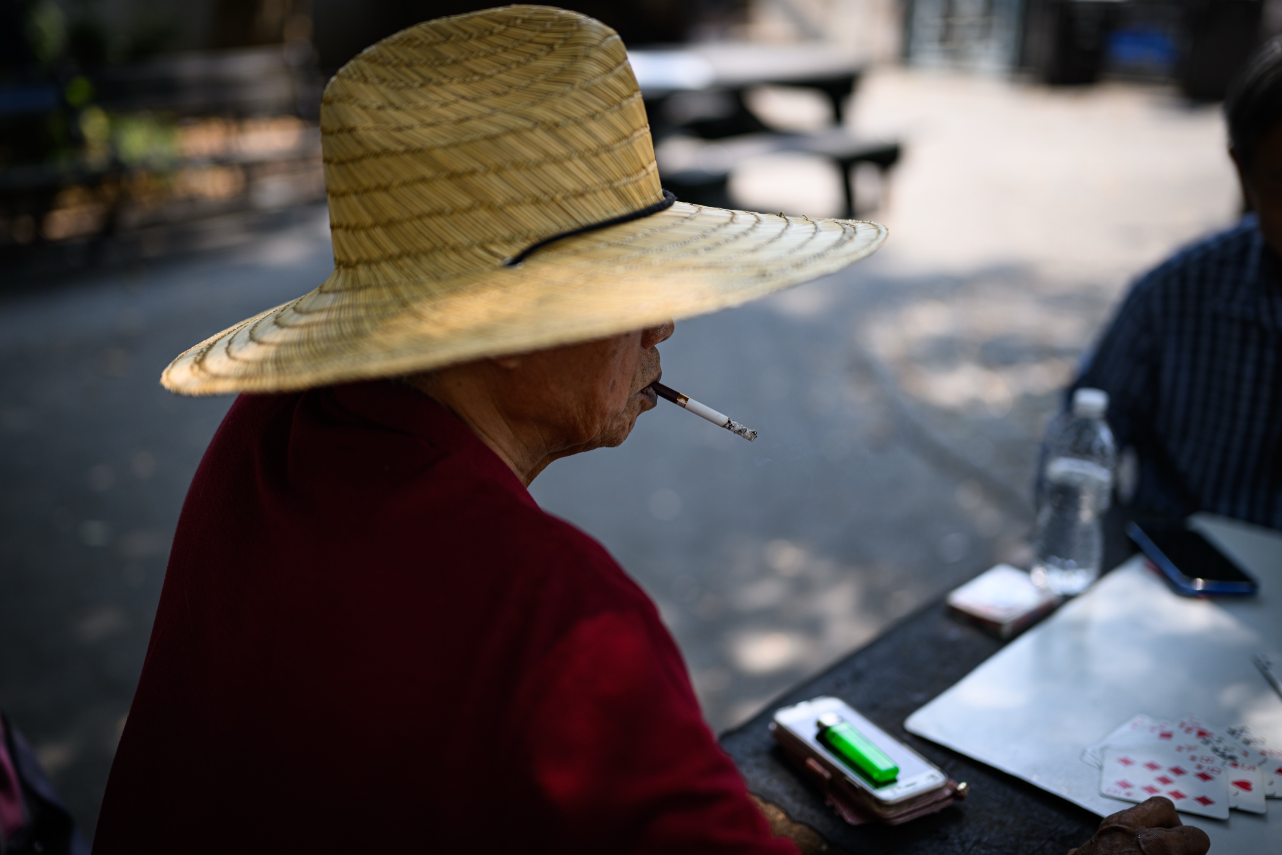 An older man smoking and playing cards under a wide straw hat